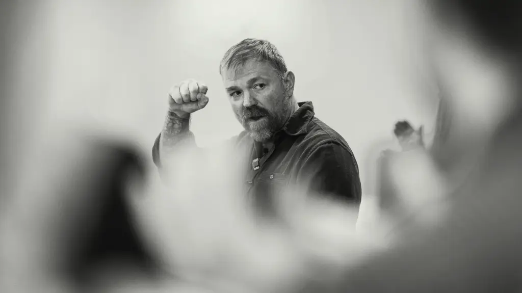 A man with light hair, a beard, and a tattooed forearm raises a clenched fist while speaking. He wears a dark, button-up shirt and is positioned against a plain, light-colored background. The image is black and white, and the scene is shot from a low angle through a crowd, with heavily blurred foreground elements framing the speaker.