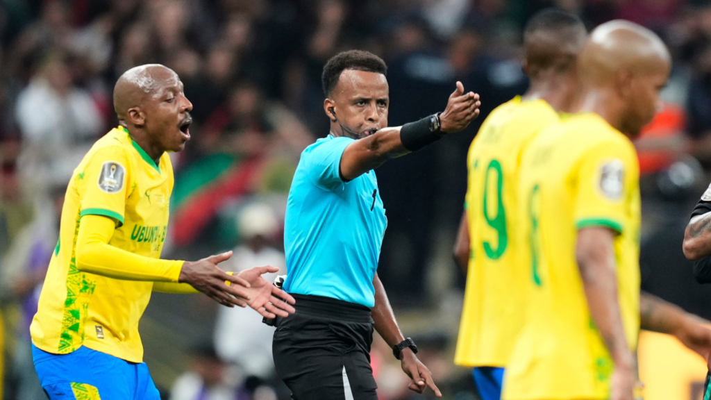 A soccer referee blows his whistle and points forward with his right hand, signaling a penalty. One player behind the referee, wearing a yellow jersey, protests the decision.