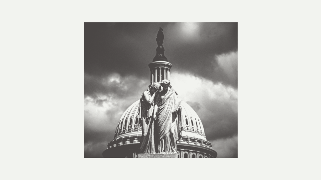 A black-and-white photo of the Peace Monument in front of the US Capitol dome. The monument is a statue of one person weeping on the shoulder of another person.