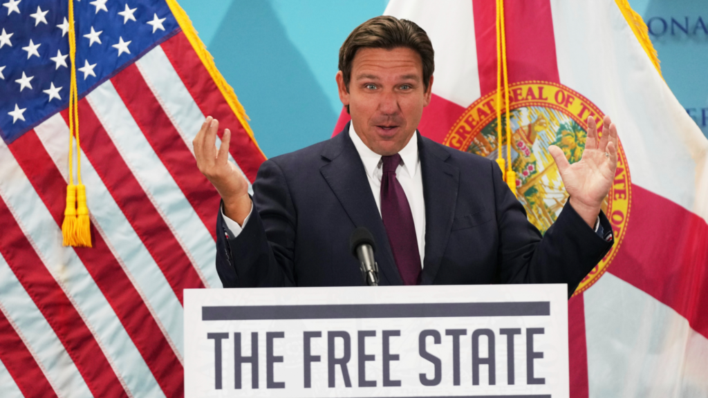 Ron DeSantis stands at a podium with a microphone as he gives a speech. Both of his hands are outstretched to emphasize his point. A sign on the podium reads