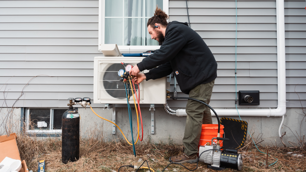 white dude with man-bun and black hoodie and some equipment installing a heat pump outside a house with light gray siding