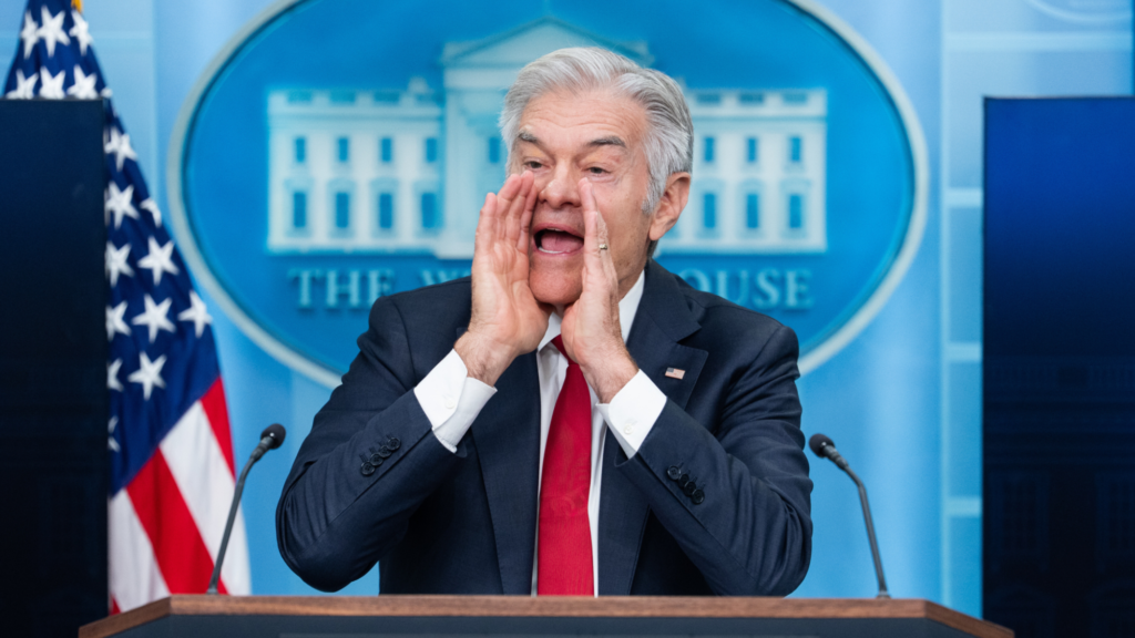 Dr. Mehmet Oz is wearing a dark blue suit and red tie. Both of his hands are cupped to his mouth to direct his voice during a press conference. He is standing at a podium with microphones.