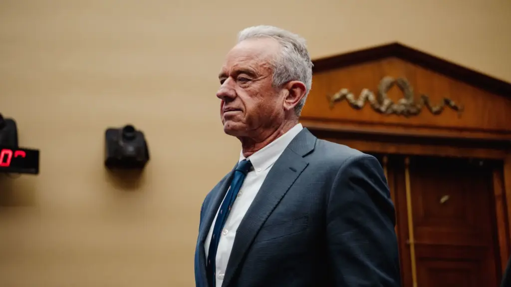 RFK Jr standing in a suit with a yellow wall behind him