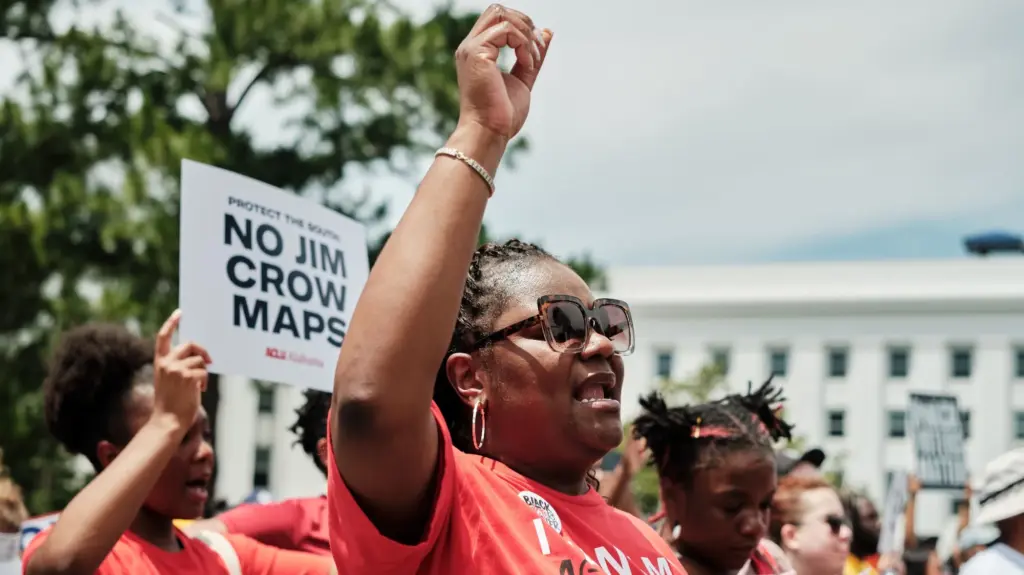 A group of black women wearing red shirts demonstrate outside a white building. One, in sunglasses, holds her right hand aloft. Another carries a sign reading 