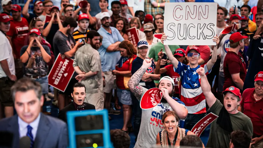 Out of focus in the foreground, a male reporter in a suit and tie speaks with a microphone in hand. Behind him, a crowd of people, many wearing red MAGA baseball caps, appear to jeer and boo. One person holds up a handwritten sign that reads “CNN sucks.”