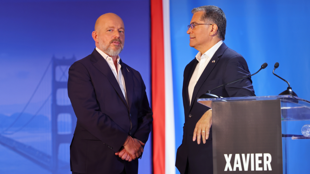 Two men in suits speak to each other, one leans on a podium with microphones attached.