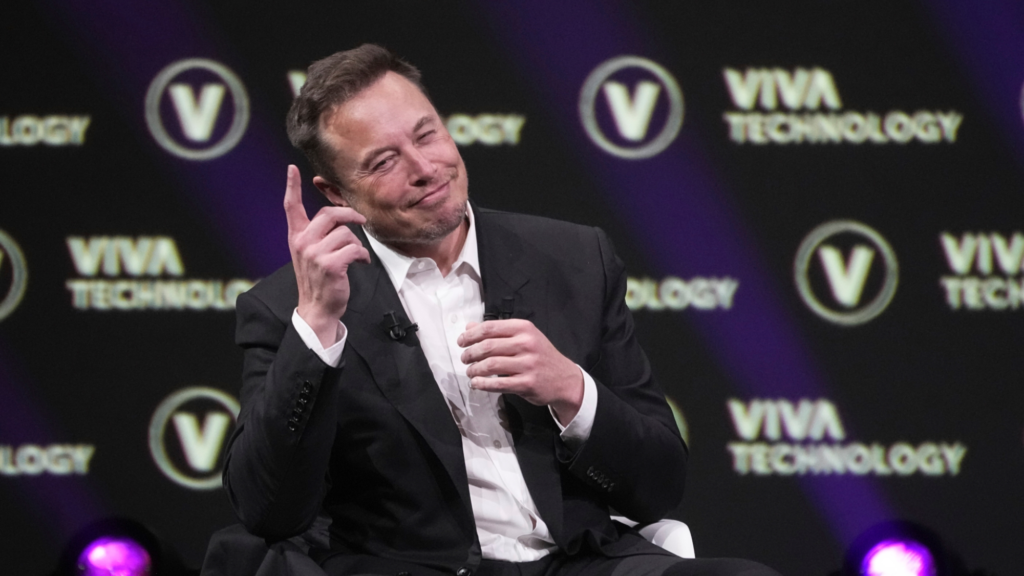 Elon Musk points up in the air with his right hand is smiling wryly while sitting in a white chair.