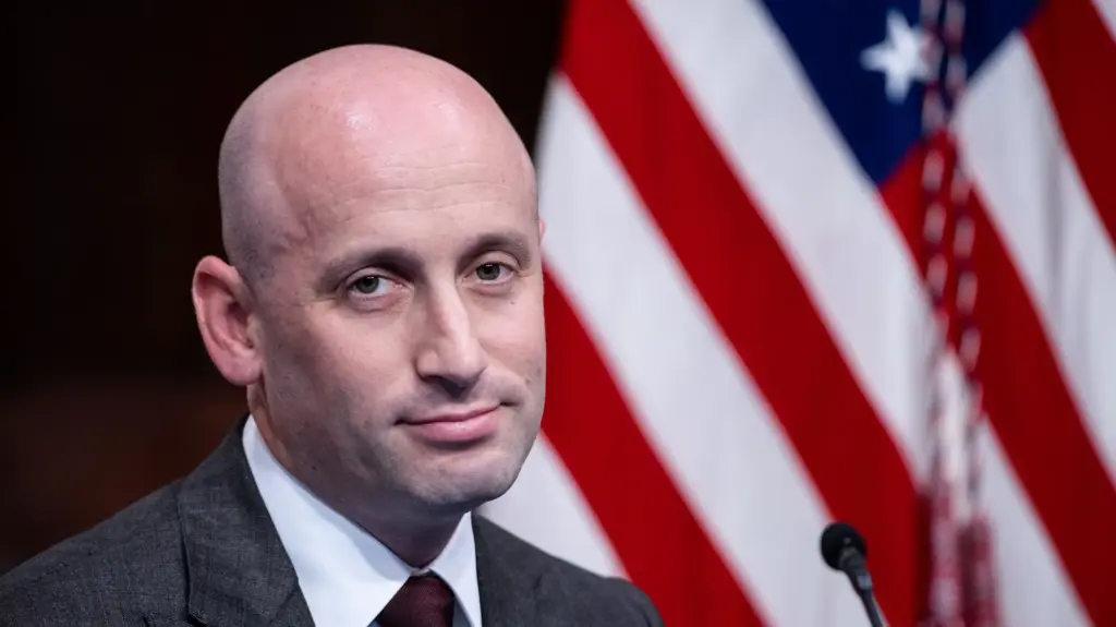 Stephen Miller, a white man, with an American flag behind him, looking very smug.