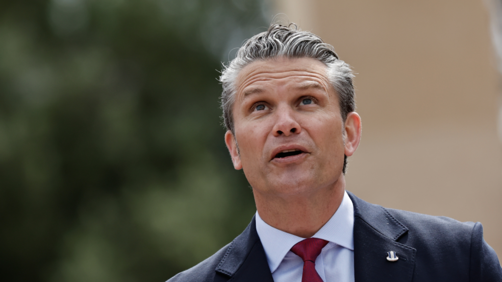 Pete Hegseth is wearing a dark suit and red tie. He is look upward as he delivers a speech outside.