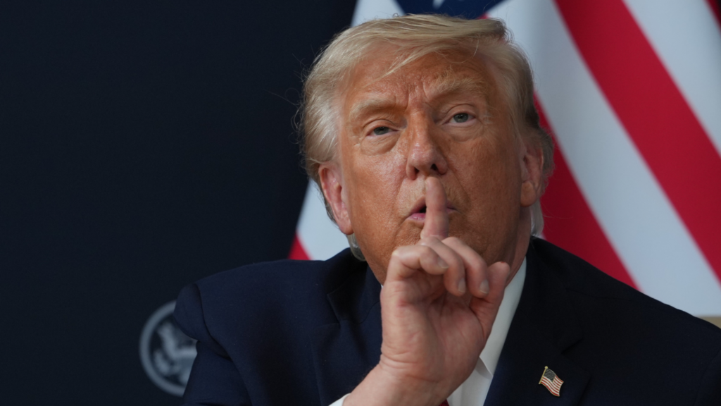 US President Donald Trump's head, shoulders, and upper chest are shown in the photo. He is holding his right pointer finger to his mouth in a shooshing motion.