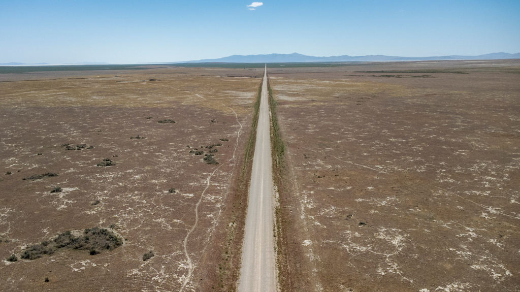 A long, straight, two-lane highway stretches into a hazy blue horizon, with flat, brown, parched land on either side as far as the eye can see.