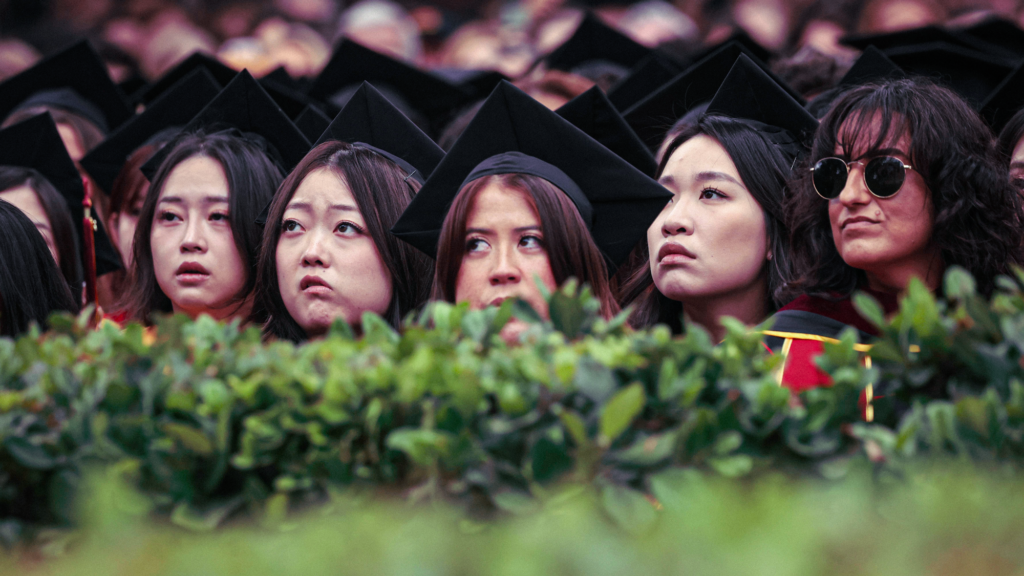 A photograph of a diverse group of students in graduation gowns and caps who are looking ahead with rapt attention. Even though seen from behind a hedge of bushes, some of the faces appear to look skeptical.