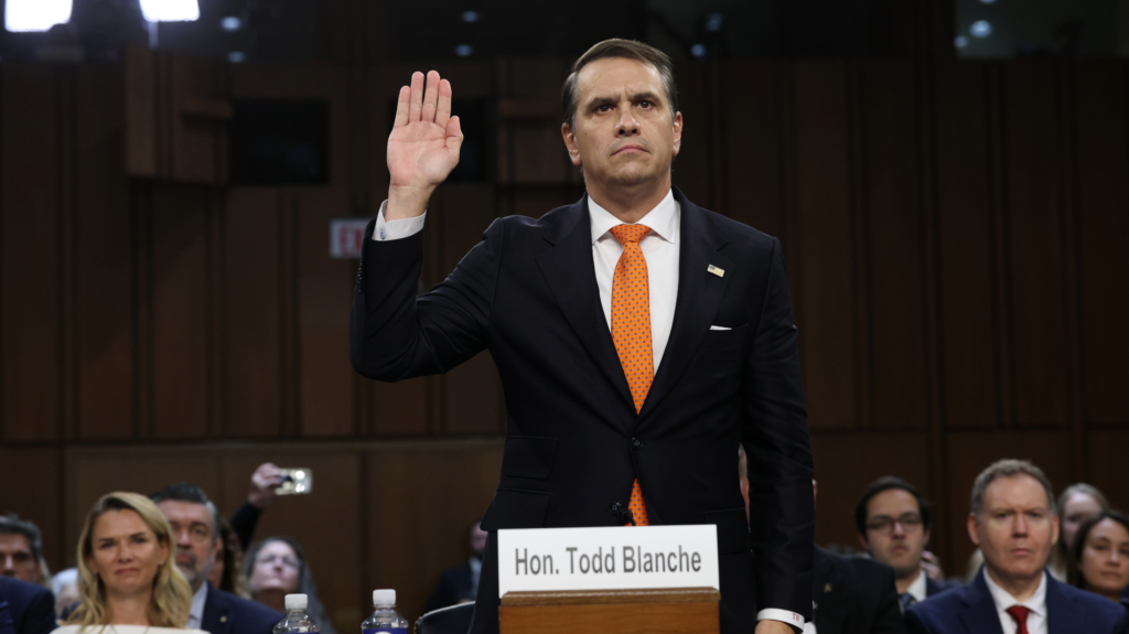 A man in a black suit raises his right hand as people seated behind him look ahead.