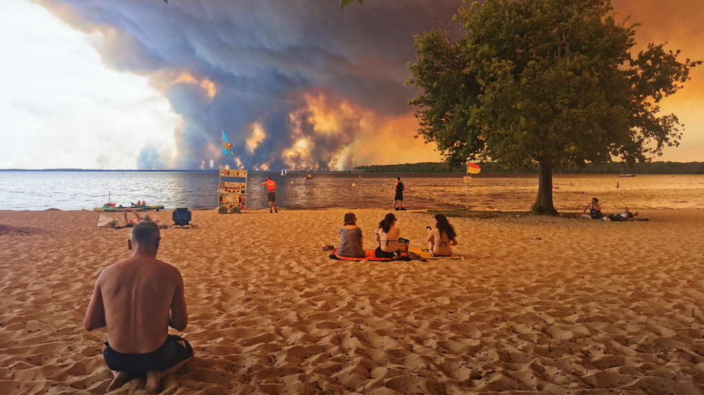 People sit on the sand at a beach in France, while clouds of smoke rise over the horizon from a wildfire.
