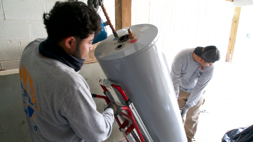 Two men move grey cylindrical gas heater.