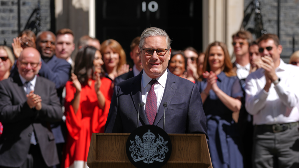 Over a dozen people in the background applaud Prime Minister Keir Starmer. Starmer is standing at a podium delivering a speech. They are all standing behind a door with the number 10 on it (10 Downing Street).