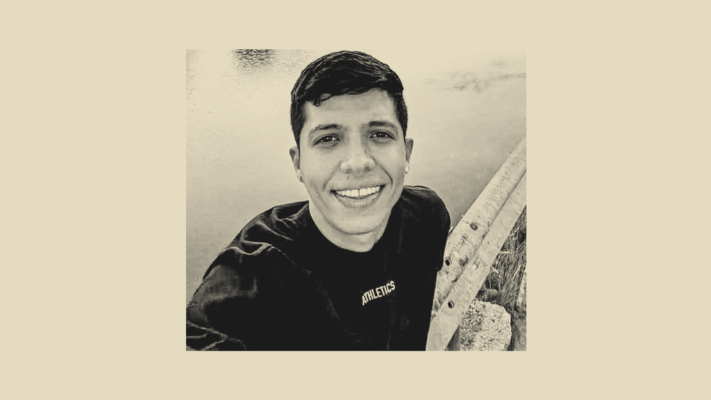 A portrait of a young man with a short hairstyle and no facial hair. Wearing a t-shirt, and offering a big smile, he looks into the camera as he sits aside a body of water. The image is overlayed with a muted gradient of black and beige.