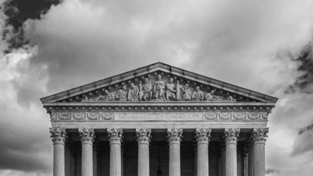 A black-and-white photo of the neoclassical facade of the United States Supreme Court building. A dark sky patchy with clouds looms overhead.