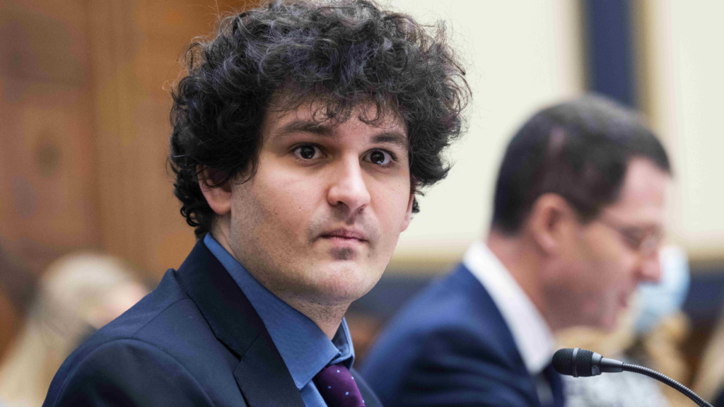 A young man with a clean-shaven face and dark curly hair wears a blue suit and shirt and purple tie with small polka dots. He’s looking toward the camera to his right as he sits before a microphone.