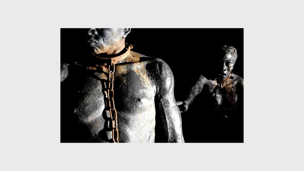 A metal sculpture of a Black man and woman starkly lit against a black background. The man looks to his right stoically, as the woman behind him reaches out to him. Both, cast in all black, have rusted metal chains fastened around their necks.
