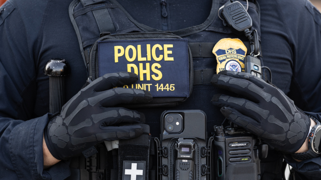 A DHS agent's chest zoomed in, with a bulletproof vest, a badge, and black-on-black skeleton gloves.