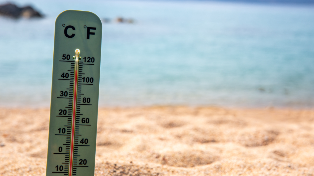 A thermometer sticking out of sand displays temperature in front of ocean.