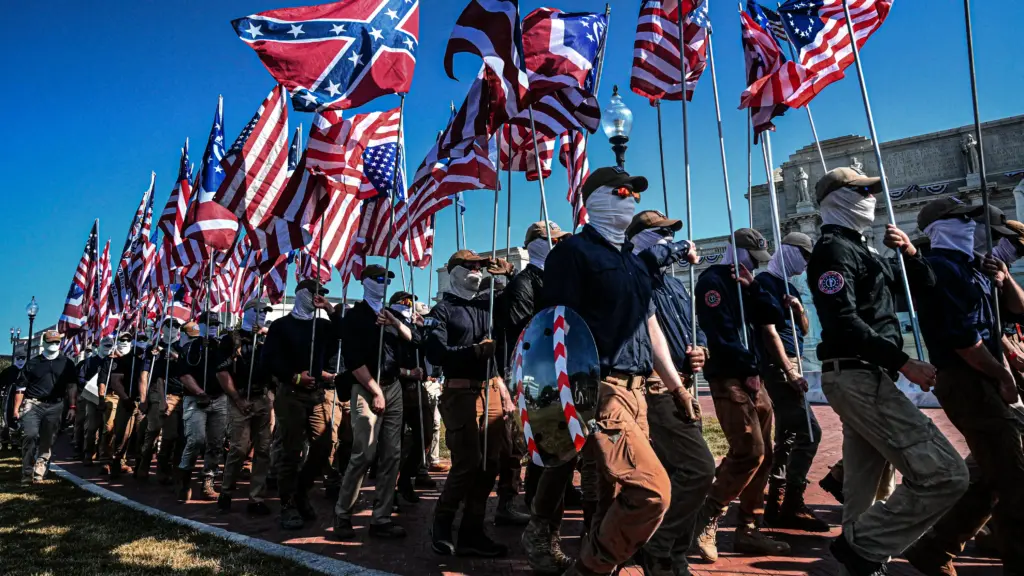 Dozens of men in blue long-sleeve shirts and brown pants gather in rows as they each hold variations of American flags. At least one carries a Confederate battle flag, and another holds a silver shield. All are wearing white face coverings, sunglasses, and baseball caps.