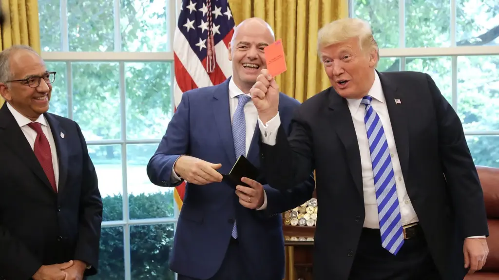 Three men in suits laugh as one with whitish blond hair holds up a red card.