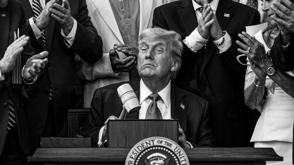 A photo of Donald Trump holding a gavel at a desk that bares the presidential seal as several people around him clap.