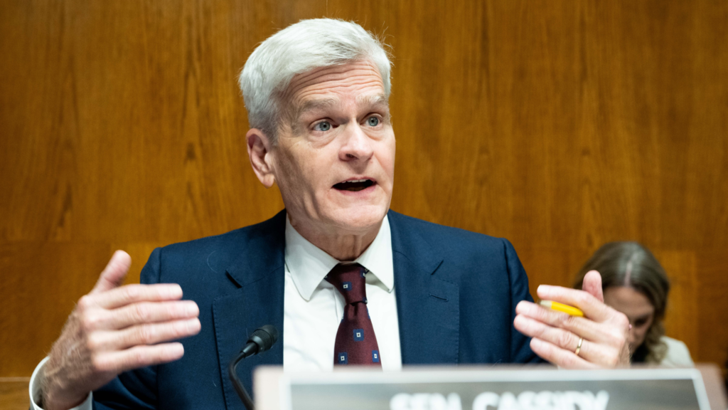 Senator Bill Cassidy, dressed in a dark suit, holds both of his hands sideways in front of himself as he speaks into a microphone. He is sitting during a Senate committee confirmation hearing. He is holding a pencil between is pointer and middle fingers in his left hand.