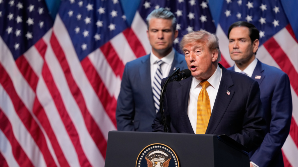 Donald Trump is shown in the center speaking into a microphone at the podium. To his right and slightly behind him is Pete Hegseth. And to Trump's left and also behind him is Marco Rubio. Several American flags are shown in the background.