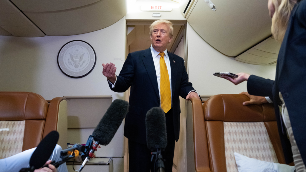 Donald Trump speaks with his his right arm outstretched in front of him. Multiple reporters are facing him holding out microphones. They are all in the Air Force One jet.