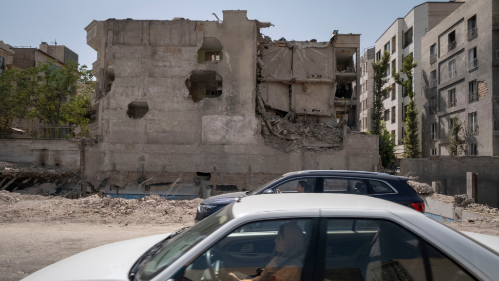 Two drivers in two different cars pass by a building destroyed by US airstrikes. Rubble from the airstrike is still on the ground.