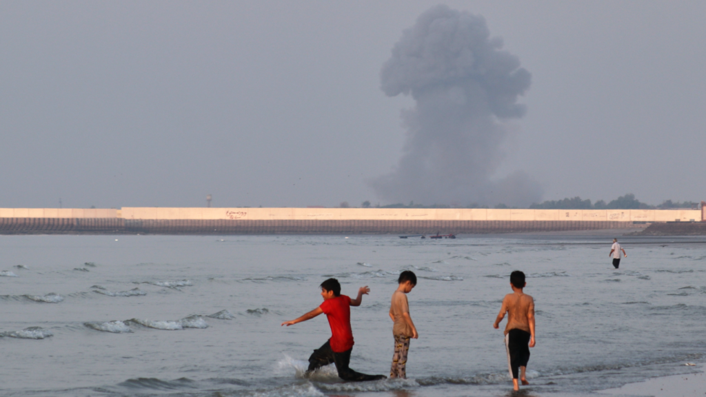 Three children are standing in shallow water. Another person is seen in the distance further into the water. In the background is a plume of smoke from an explosion.