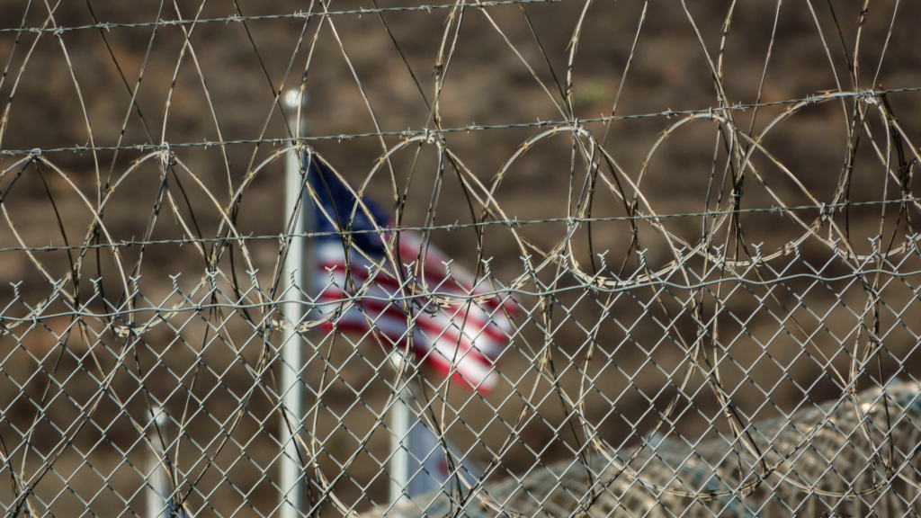 An American flag can be seen through the barbed wire surrounding the CoreCivic Otay Mesa Detention Center on October 4, 2025 in San Diego, California.