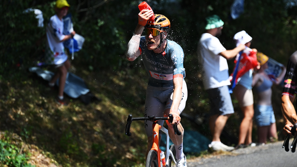 A man in an orange helmet riding an orange bike pours water onto his head from a red water bottle.