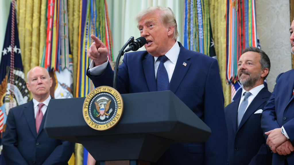 Donald Trump, white man in blue suit points as he makes speech behind podium with other men standing behind him.