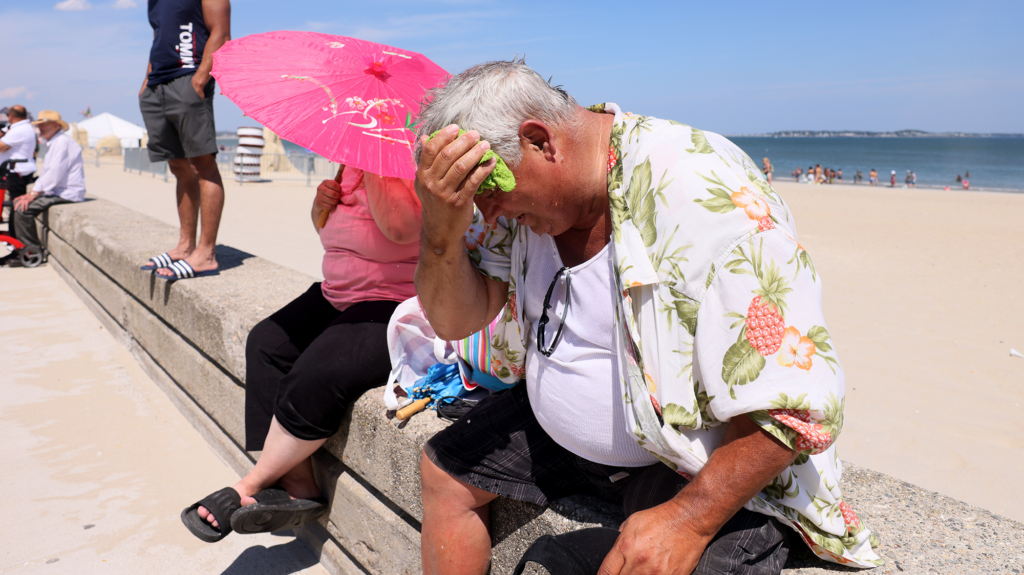 Man with white hair in Hawaiian shirt wipes the sweat off his face while sitting next to a woman holding a pink parasol at the beach.