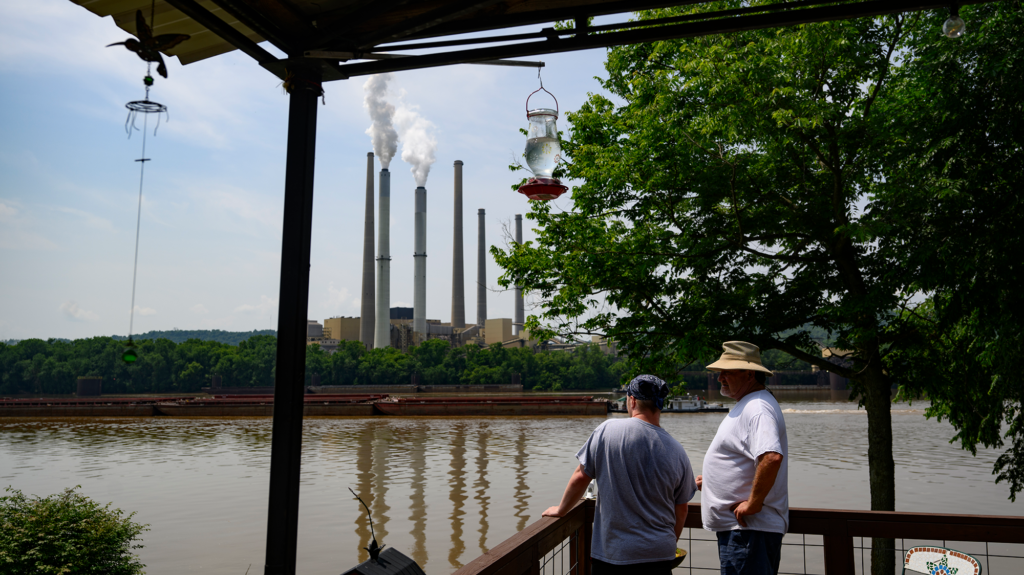 Two people stand on a wooden deck looking across a muddy river at a coal power plant releasing emissions.