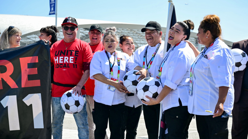 Nine workers at SoFi Stadium are seen standing next to each other. Five of them are wearing white shirts and two are wearing red union shirts. Two other people are holding a union banner in front of themselves.
