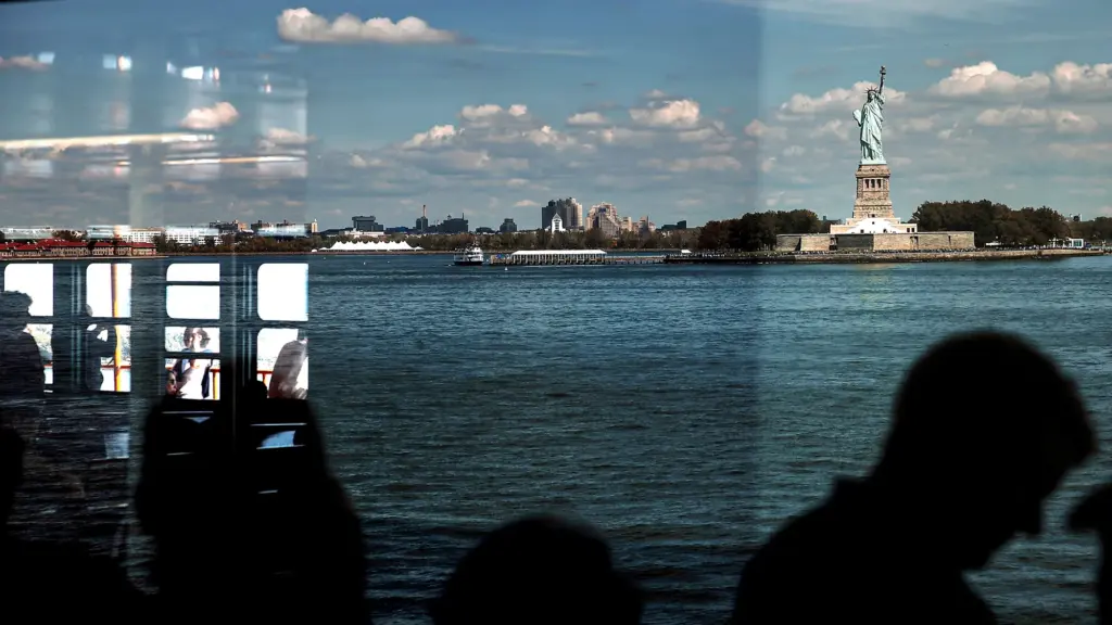 A photograph captures a scenic view of New York Harbor framed through a reflection-filled window on the Staten Island Ferry. Silhouettes of several passengers appear in the bottom half of the image, looking out toward the choppy blue water. Across the water stands the Statue of Liberty.