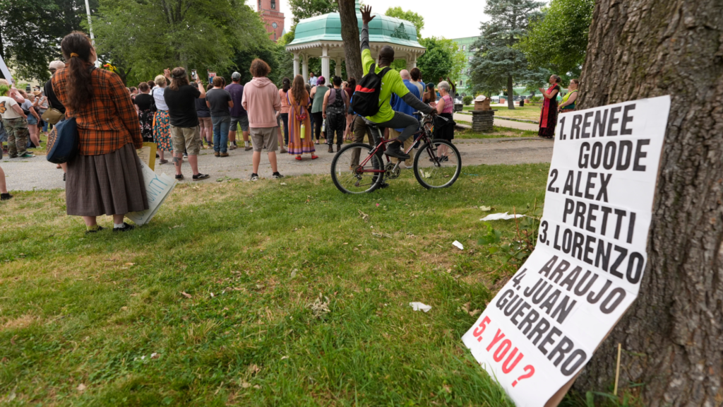 Dozens of people are shown gathered at a vigil for Johan Sebastián Durán Guerrero, who was shot and killed by ICE officers. A sign shows some people killed by federal officers: Renee Goode, Alex Pretti, Lorenzo Araujo, and Juan Guerrero.