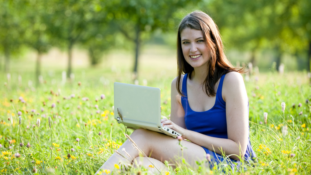 A woman sits and uses a laptop in a meadow, smiling.