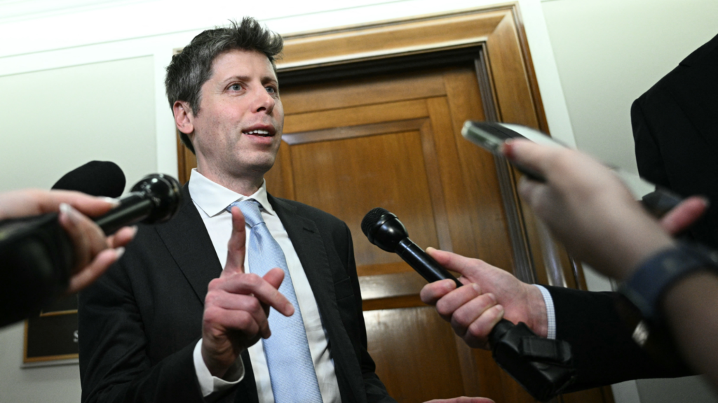 Sam Altman is wearing a suit and is speaking to multiple journalists holding out recording devices. Altman is pointing up with his right pointer finger as he speaks.