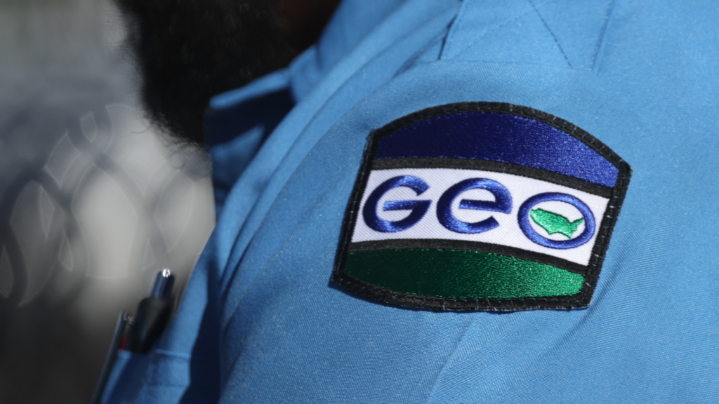 A photo of an officer's left shoulder. The officer is wearing a blue uniform containing patch with the GEO Group logo.