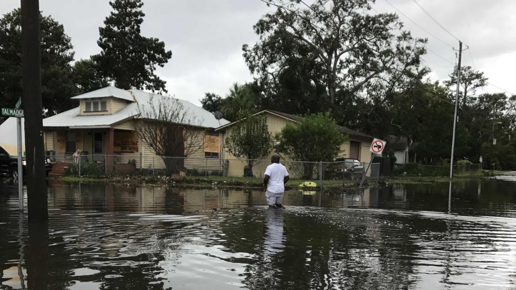A person stands in knee-deep water during a flood in front of a house.