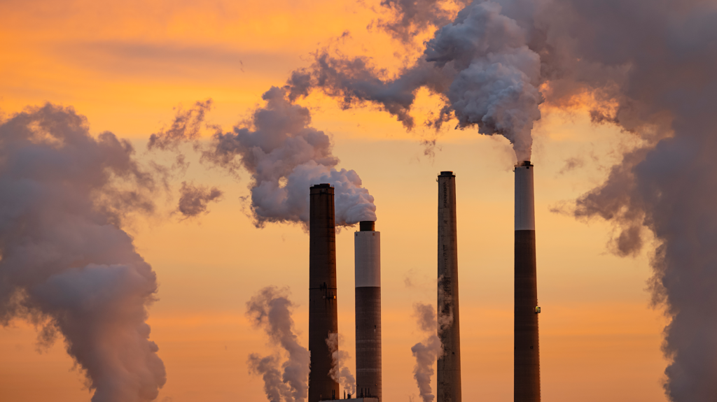 Smokestacks from a coal-fired power plant release smoke in front of an orange sky.