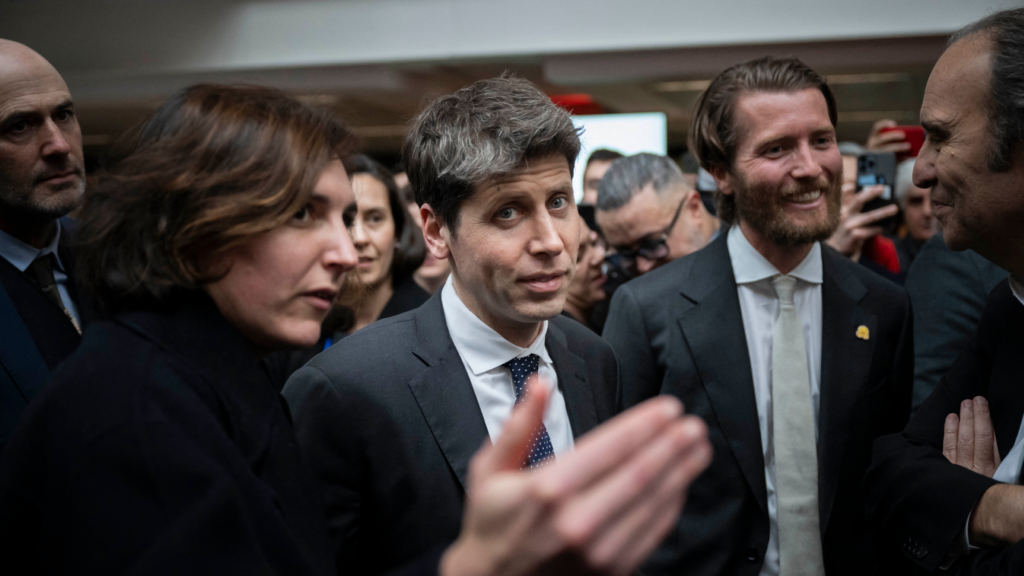 A man in a dark suit and tie stares directly into the camera as he stands among others, who are also formally dressed. A woman beside him extends her right hand as if making an introduction.