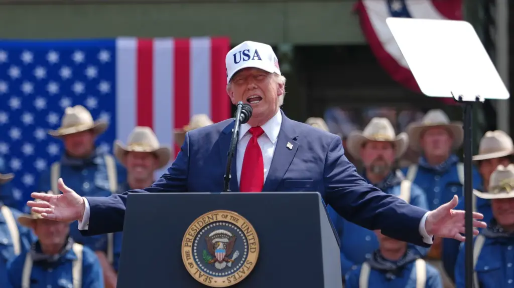 Donald Trump in a blue suit and USA baseball cap at a podium in front of men in blue shirts and cowboy-style hats.