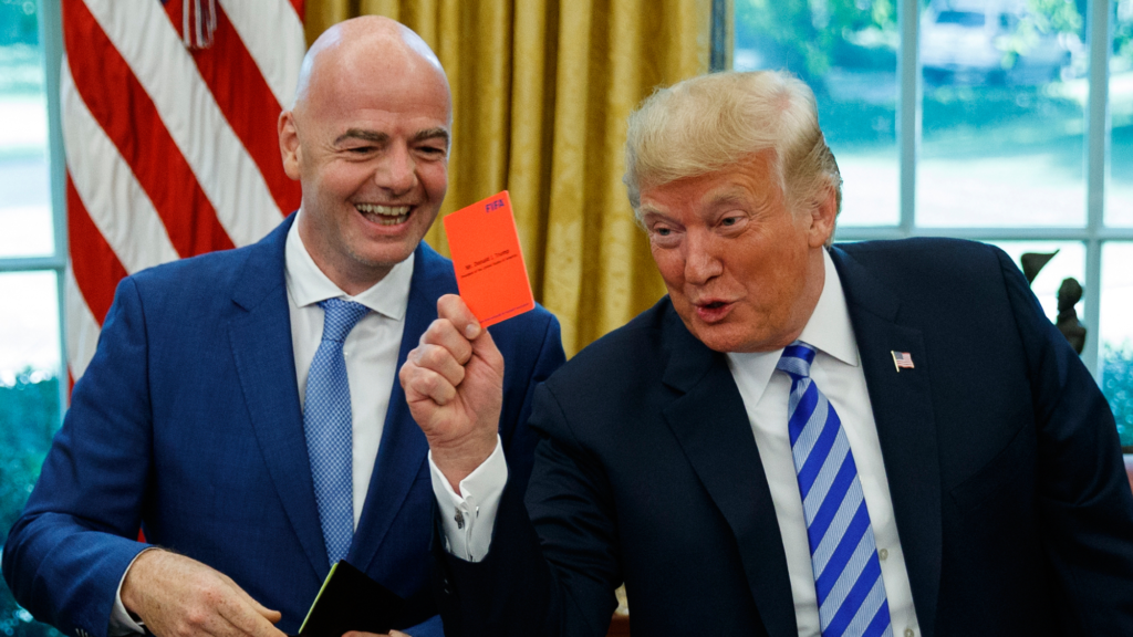 President Trump stands to the left of Gianni Infantino. Trump is holding up a red card with his right hand. Both men are smiling.